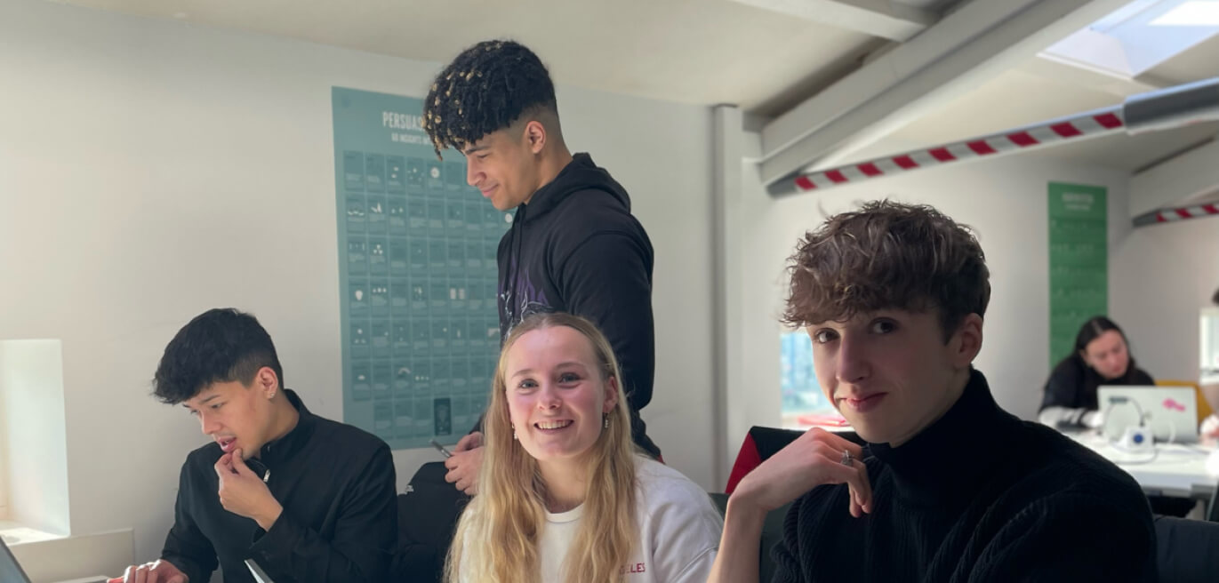 Twee studenten kijken lachend naar de camera, op de achtergrond zijn twee studenten aan het werk.